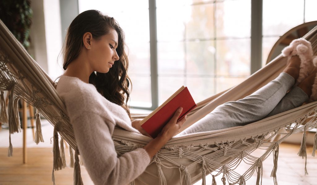 Dark-haired girl dressed in pants, sweater and warm slippers reads a book lying in a hammock